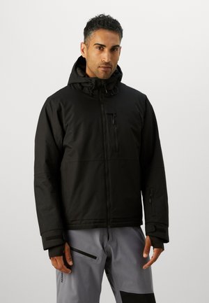 Man in zwart hooded jack met ritssluiting op de borst en grijze broek met ritssluiting op de dij, staande tegen een effen achtergrond.