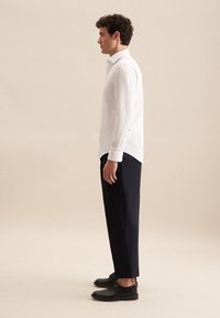 Biała koszula z długim rękawem i kołnierzykiem, noszona z granatowymi spodniami i czarnymi butami. Materiał wydaje się gładki z bocznymi rozcięciami w pobliżu dołu.