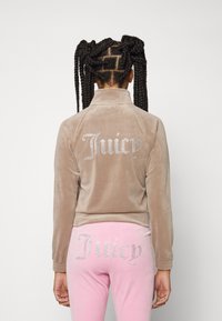 Velours-Trainingsanzug in hellbraun mit silbernen Strasssteinen "Juicy" auf dem Rücken der Jacke und dem Gesäß der rosa Hose. Strukturierter, taillierter Schnitt.