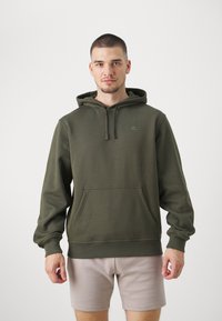 Felpa con cappuccio verde oliva in morbido cotone, dotata di tasca a marsupio frontale e cordino, abbinata a pantaloni corti beige chiaro.