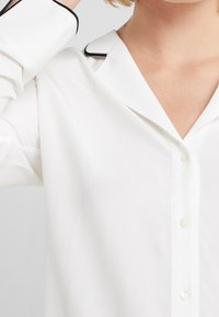 Witte button-up shirt met een zachte textuur, voorzien van een klassieke kraag en zwarte pipingaccenten langs de kraag en manchetten.