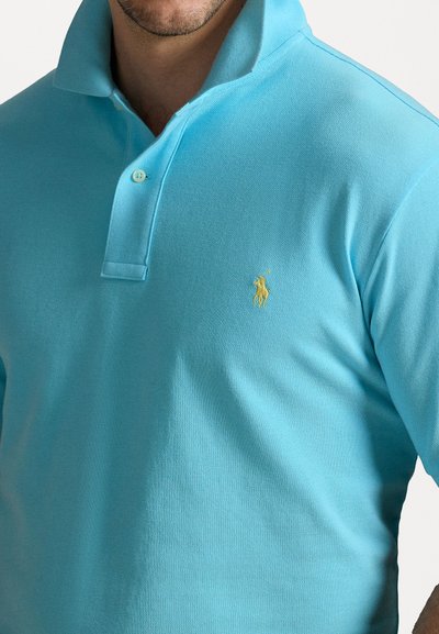 Ανοιχτό μπλε πόλο μπλούζα φτιαγμένη από υφασμάτινη υφή, με κολάρο τριών κουμπιών και ένα μικ�ρό κίτρινο λογότυπο στο αριστερό στήθος.