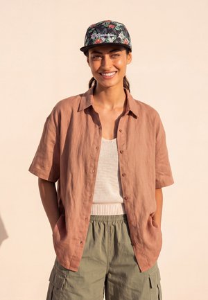 Hymyilevä nainen, jolla on kukallinen lakki, löysä ruskea lyhythihainen paita, valkoinen neulepaita ja vihreät cargo-housut, kädet taskuissa.