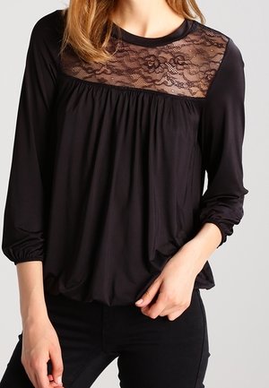 Femme portant un chemisier à manches longues noir avec un yoke en dentelle florale et des détails froncés, associé à un pantalon noir.