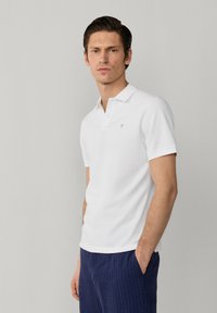 Uomo che indossa una polo bianca a maniche corte e pantaloni blu a righe sottili, in piedi con una mano in tasca davanti a uno sfondo grigio uniforme.