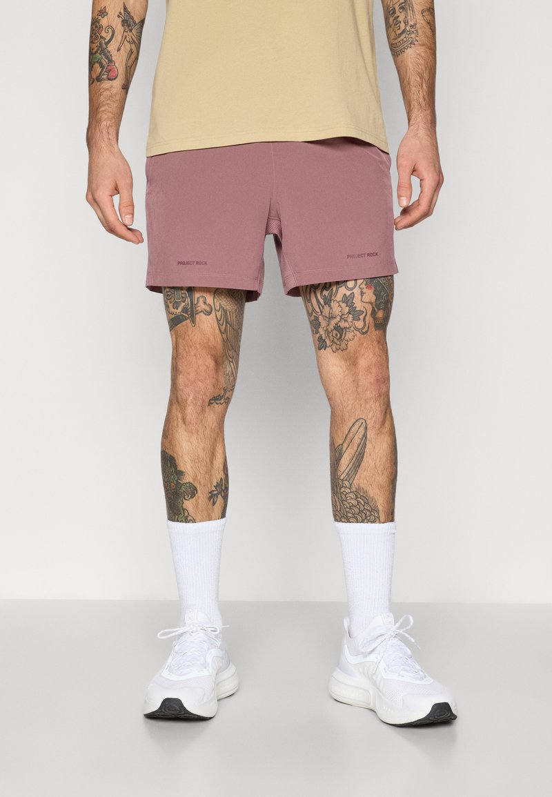 Kurze sportliche Herren-Shorts in staubiger Rose, aus leichtem Material, mit elastischem Bund und Logo-Details. Getragen mit weißen Knöchelsocken und Sneakers.
