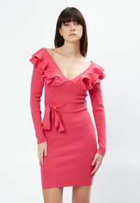 Abito rosa a coste con scollatura a V profonda, dettagli arricciati sulle spalle e vita legata. Maniche lunghe e silhouette aderente.