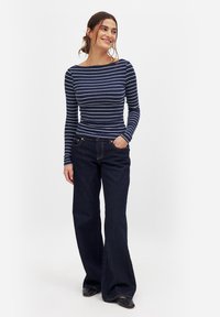 Mornarsko modra in bela črtasta majica z dolgimi rokavi, kombinirana s temno flared denim hlačami. Majica ima čolnasti izrez in rebrasto teksturo.