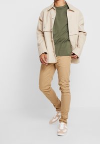 Giacca beige con bottoni, t-shirt verde oliva, pantaloni aderenti color sabbia e sneakers beige con dettagli bianchi. Outfit moderno e casual.