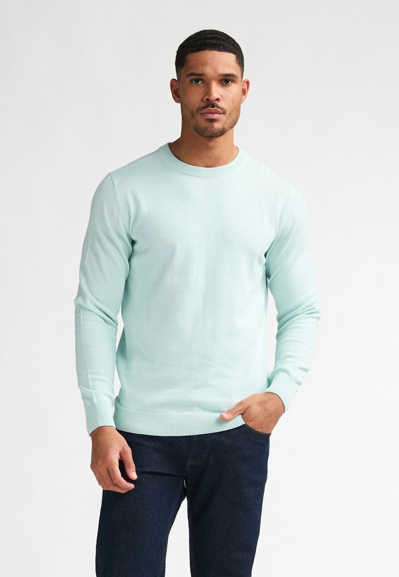 Sweter z dzianiny w jasnym miętowym kolorze, z długimi rękawami, okrągłym dekoltem oraz ściągaczami na mankietach i dole, zestawiony z ciemnymi dżinsami. Gładka faktura.