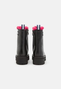 Botas de cuero negro a nivel del tobillo con suelas gruesas, forro de piel de oveja rosa en la parte superior y lengüetas de tirón azul y blanca en la parte posterior.
