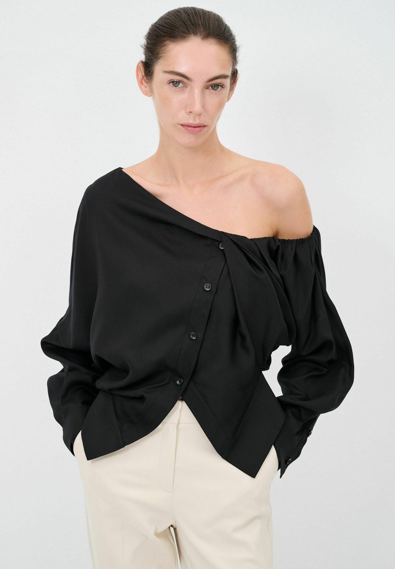 Blusa nera off-the-shoulder con bottoni sul davanti, tessuto drappeggiato e orlo diagonale, abbinata a pantaloni chiari.