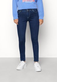 Jeans skinny azul oscuro con una textura suave, diseño de cinco bolsillos y costuras amarillas en contraste, combinados con zapatillas blancas.