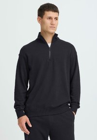 Jeune homme portant un pull noir à demi-fermeture éclair et un pantalon noir assorti, debout avec une main dans la poche devant un fond blanc.