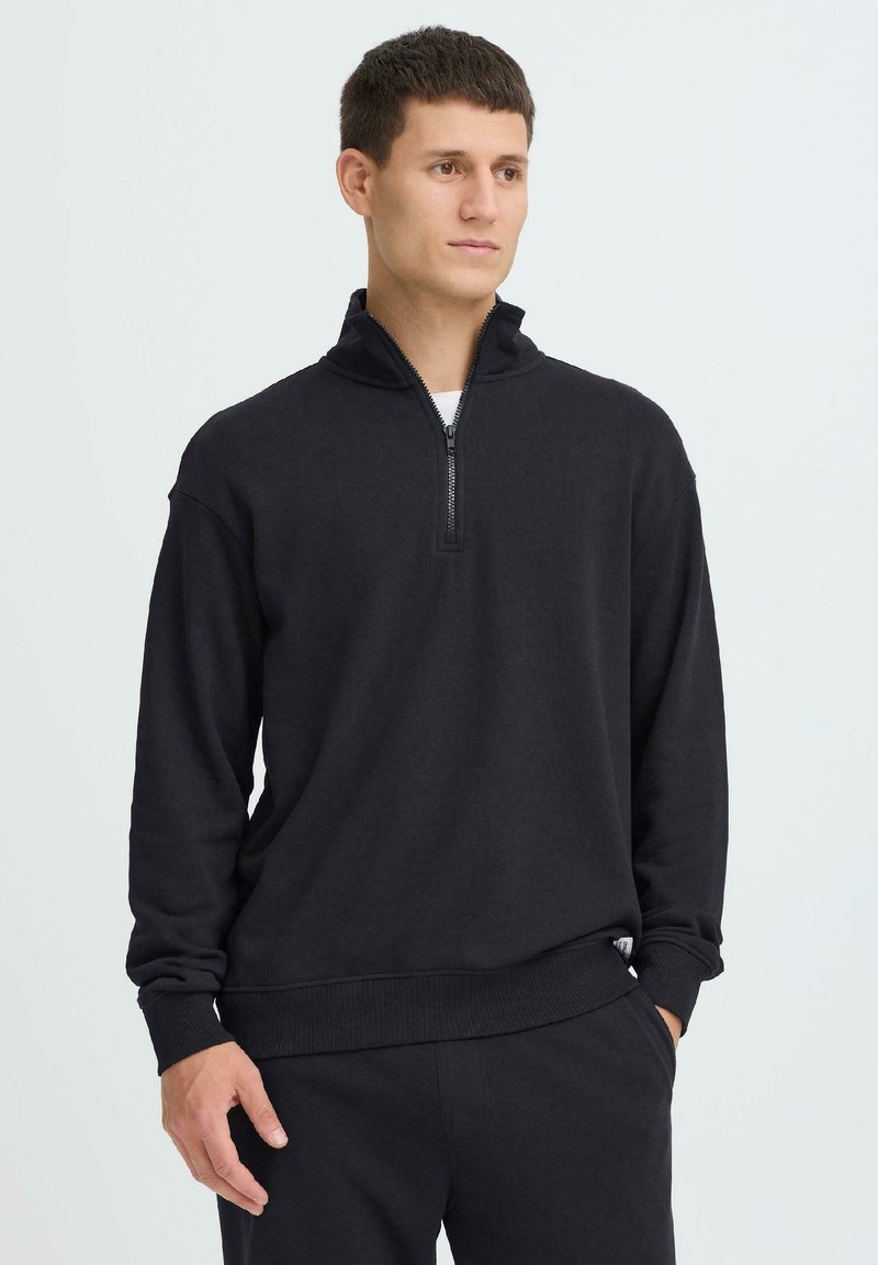 Jeune homme portant un pull noir à demi-fermeture éclair et un pantalon noir assorti, debout avec une main dans la poche devant un fond blanc.