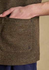 Brown herringbone patterned fabric with a visible pocket and stitched accents. Features a branded logo in the lower right corner.