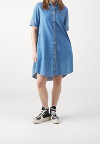 Abito in denim azzurro chiaro con maniche corte, chiusura con bottoni sul davanti e tasca sul petto. Presenta un orlo curvo ed è indossato con sneakers alte nere.