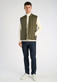 Olivgrünes ärmelloses Weste mit gestepptem Muster, getragen über einem cremefarbenen Rollkragenpullover. Dunkelblaue Jeans und weiße Sneakers runden das Outfit ab.