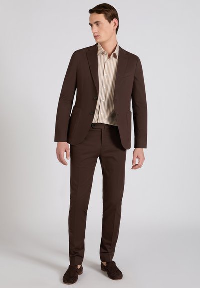 Man draagt een donkerbruin pak met een colbert met twee knopen, een beige gestreept overhemd, bijpassende broek en donkerbruine loafers, staand tegen een effen achtergrond.