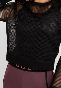 Czarny siatkowy top crop z teksturowanym wzorem, z solidnym czarnym dekoltem i rękawami. Logo nadrukowane w kolorze ton w ton.