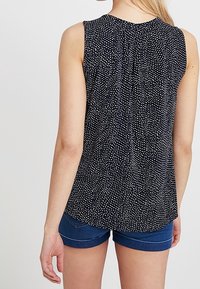 Bakvy av en ärmlös svart blus med vita prickar, som har en rundad fåll och subtila veck vid halsen. Kombinerad med blå shorts.