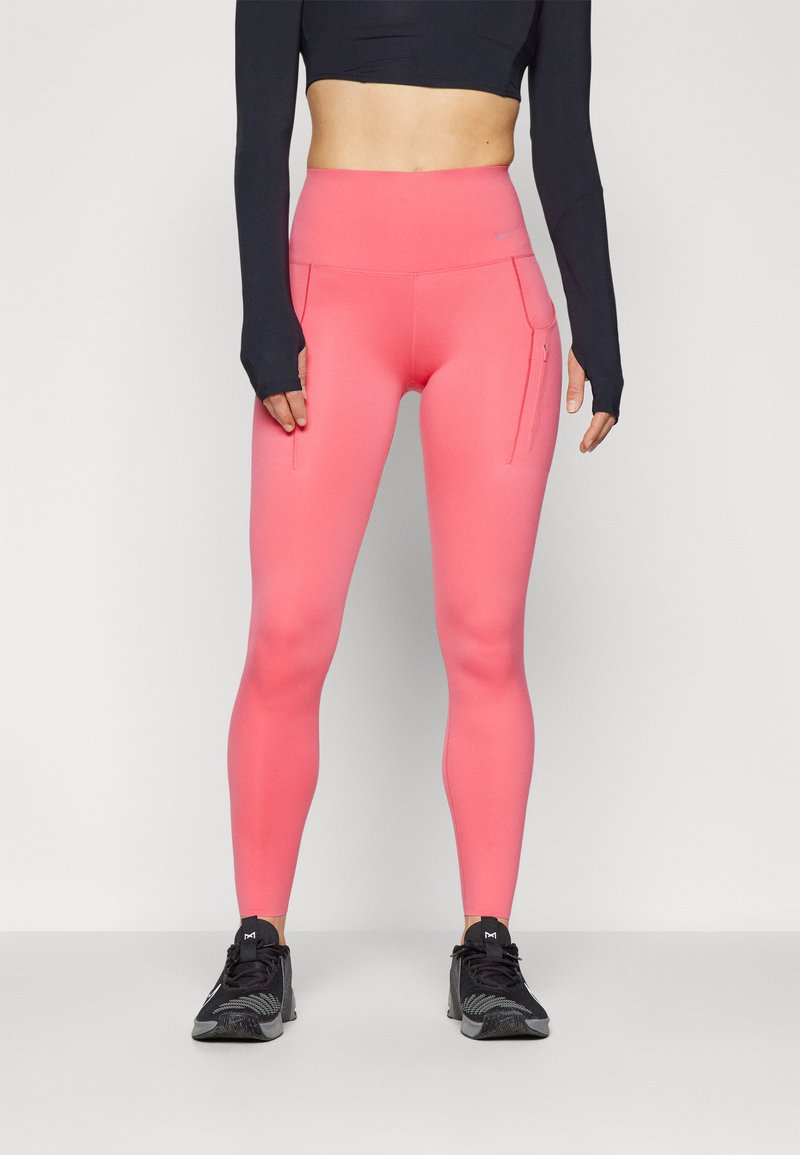 Leggings taille haute, rose vif, avec poches latérales, fabriqués en tissu extensible. Associés à des chaussures de sport noires et à un haut à manches longues.