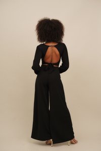 Black jumpsuit with a deep back cutout, long sleeves, and a tie waist. Smooth fabric, wide legs, and a visible tattoo along the spine.
