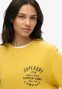 Żółta bluza z okrągłym dekoltem, ściągaczami na rękawach i teksturowanym materiałem. Branding "Superdry" w czerni na piersi.