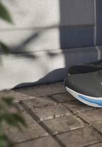 Scarpe da ginnastica nere con tomaia texturizzata, suola bianca e dettagli blu, posizionate su una superficie in mattoni con ombre morbide.