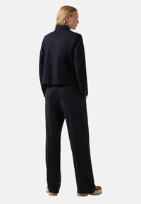 Woman with blonde hair wearing a navy jacket and matching wide-leg pants, standing with hands in pockets, facing away against a plain background.