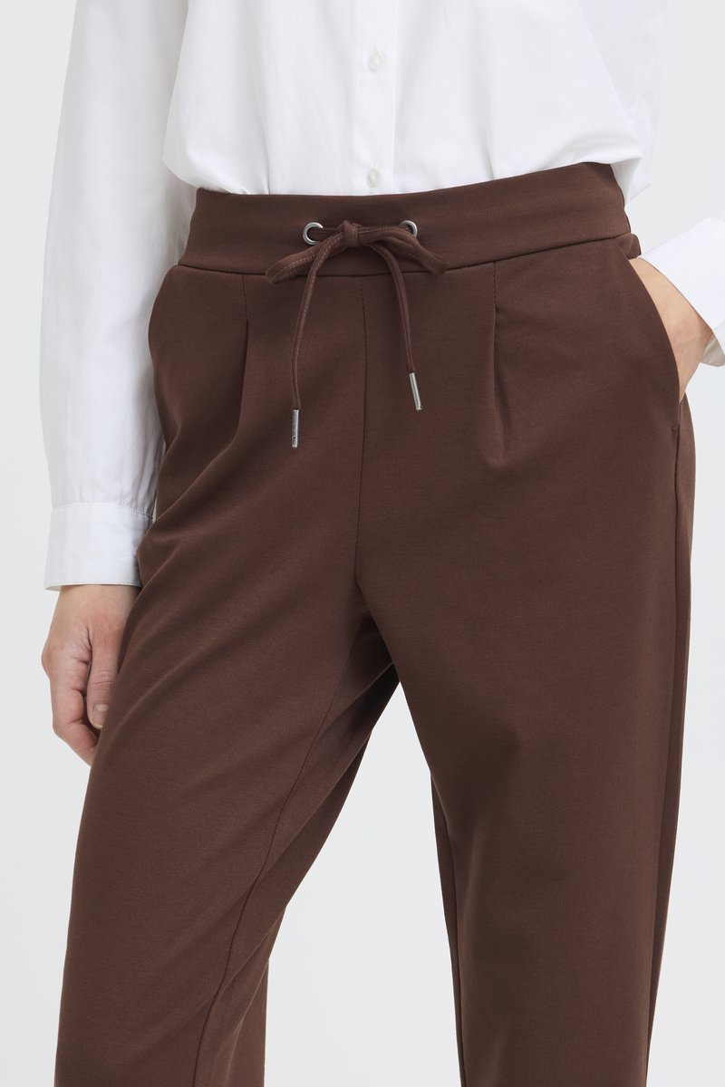 Frau trägt braune, hochtaillierte Hose mit Kordelzug und weißes Hemd mit Knöpfen, Hand in der Hosentasche, steht vor einem einfarbigen Hintergrund.