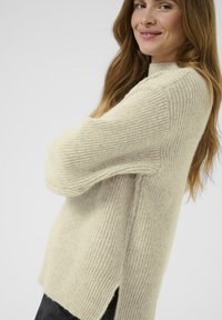 Woman with long brown hair wearing a beige knitted sweater with a side slit, smiling softly, with arms crossed.