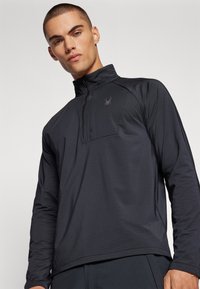 Schwarzer Pullover mit langen Ärmeln und einem Viertel-Reißverschluss, ausgestattet mit einem dezenten Strukturmuster und einem kleinen Logo auf der Brust. Glatte Stoffoberfläche.