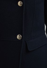 Mørkeblå uldfrakke med glat tekstur, guldmetalliske knapper og et skræddersyet design med sidelommer.