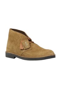 Bottines désert en suede marron avec un bout arrondi, fermeture à lacets et semelle en caoutchouc. Présente une étiquette avec le logo "Clarks" accroché.