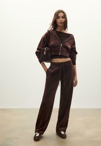 Brązowy dres z weluru, składający się z krótkiej kurtki zapinanej na zamek i luźnych spodni, dopełniony małą torebką o fakturowej powierzchni oraz sneakersami.