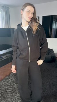 Schwarzer Kapuzenpullover mit Reißverschluss, Taschen und Logo auf der Brust, kombiniert mit locker sitzender schwarzer Jogginghose, beide aus weichem Stoff.