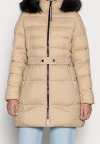 Béžová prošívaná bunda s černým zipem a páskem, s kapucí z umělé kožešiny. Prošívaná textura s logem na hrudi a žebrovanými manžetami.
