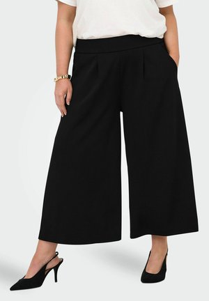 Žena má na sebe čierne široké skrátené nohavice, biely vsadený top, čierne slingback lodičky a zlatý náramok, stojí s jednou rukou v vrecku.