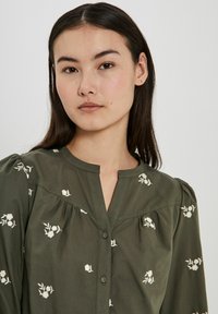 Olivgrön blus med blommig broderi, knäppning fram och plisserade detaljer vid axlarna. Lätt bomullsmaterial, avslappnad passform.