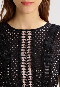 Kvinna klädd i en svart spetsklänning med intrikata mönster och genomskinliga detaljer över lätt tyg på bröstet och axlarna.