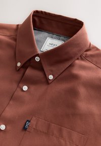 Chemise marron à col avec des boutons blancs, une poche poitrine et une étiquette portant la mention "NEXT". Tissu lisse, coupe ajustée et intérieur du col contrastant.