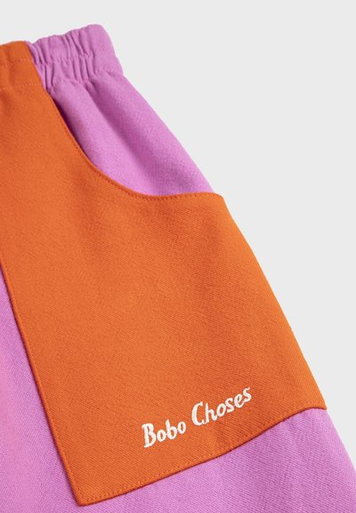 Gros plan sur un tissu montrant un vêtement rose avec une poche appliquée orange brodée de "Bobo Choses" en fil blanc.
