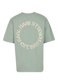 Lys grønn oversized t-skjorte med et sirkulært kremfarget grafisk tekstdesign på baksiden, med "KARL KANI STUDIOS EST. 1989."