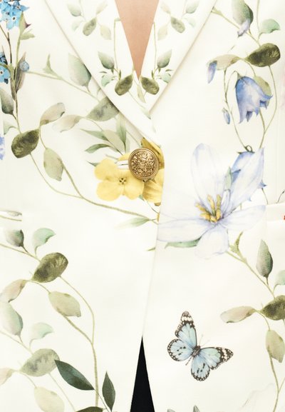 Λευκό σακάκι με πράσινο φύλλο και μπλε λουλούδι τυπωμένα, χρυσό διακοσμητικό κουμπί και μια μπλε πεταλούδα κοντά στο κάτω δεξιό μέρος.