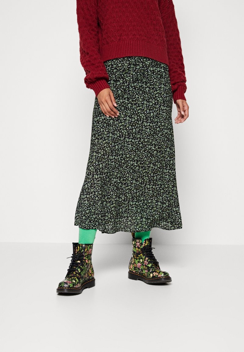 Personne portant un pull rouge, une jupe midi noire fleurie, des chaussettes vert vif et des bottines noires à fleurs lacées, debout devant un fond uni.