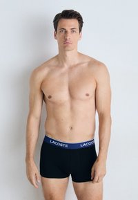 Boxer shorts negros con una cinturilla azul que presenta el logo de Lacoste, hechos de una tela suave, llevados por un hombre frente a un fondo claro.