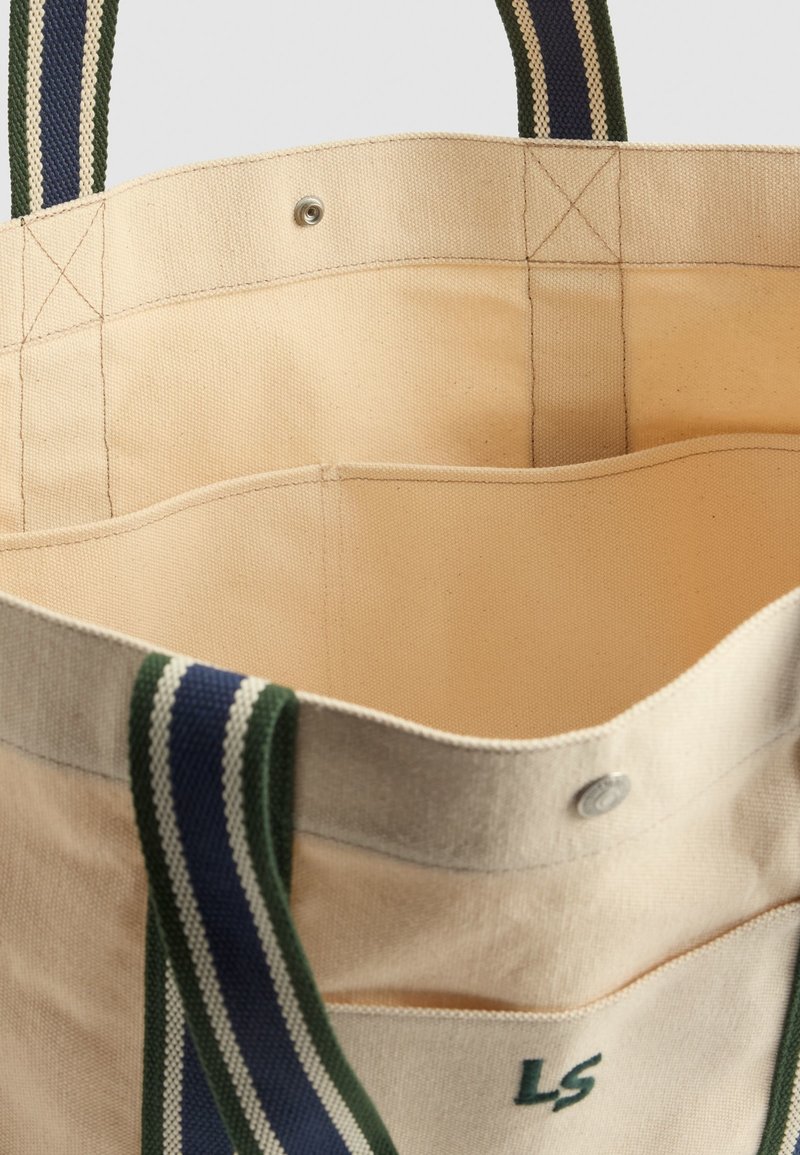 Sac fourre-tout en toile beige avec des poignées rayées vertes et bleu marine, des poches intérieures et une fermeture à bouton-pression. Dispose de détails de couture simples.