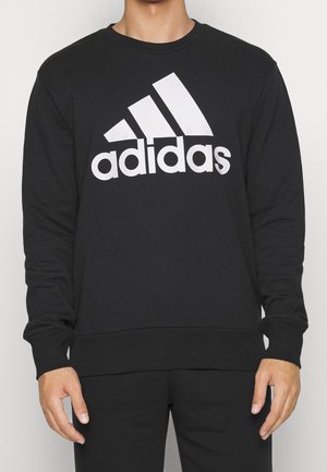 Mężczyzna w czarnej bluzie z długim rękawem z dużym białym logo Adidas na piersi, stoi na jednolitym tle.