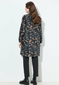 Femme avec de longs cheveux ondulés portant une tunique à manches longues à motif floral sombre, un pantalon noir et des bottes noires à la cheville, standing face à un mur blanc.